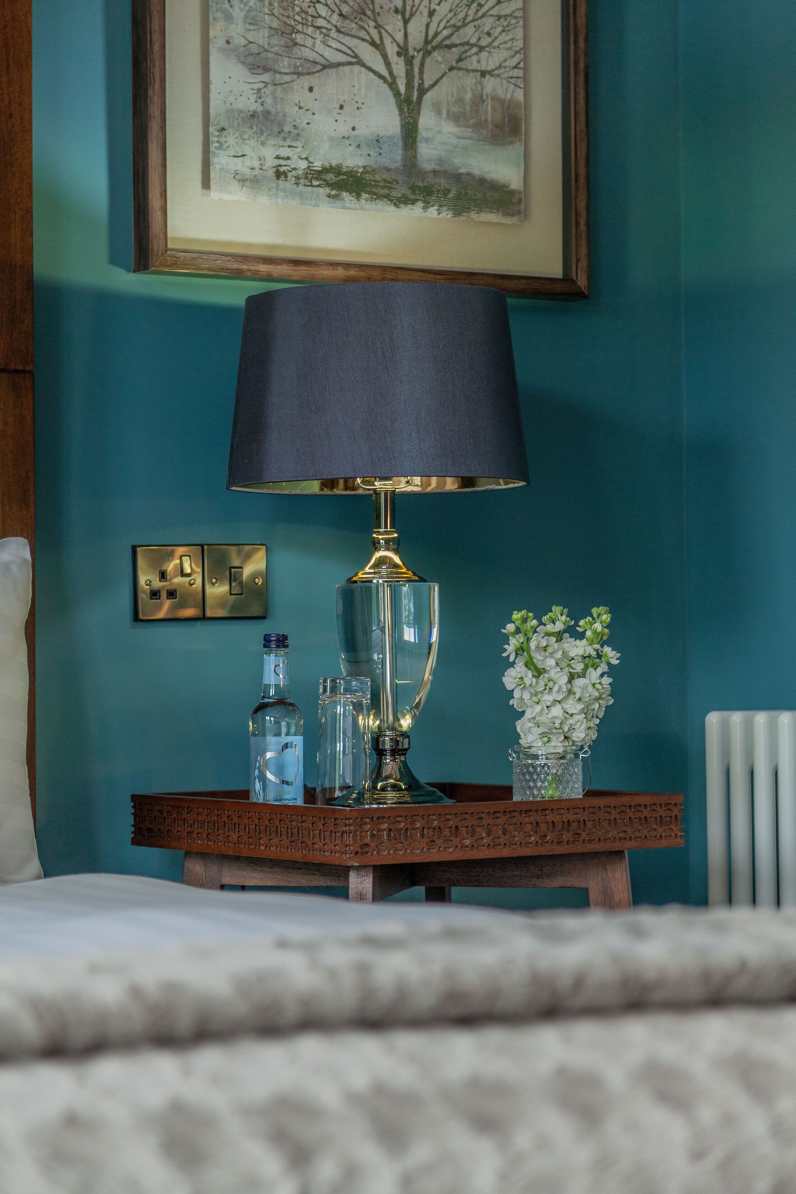 A bedside table with a lamp, a vase with flowers, and a framed picture on teal walls.