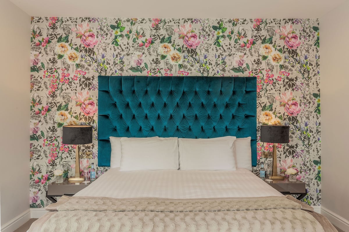 A bedroom with a floral wallpaper, a teal upholstered headboard, and two bedside tables with lamps.