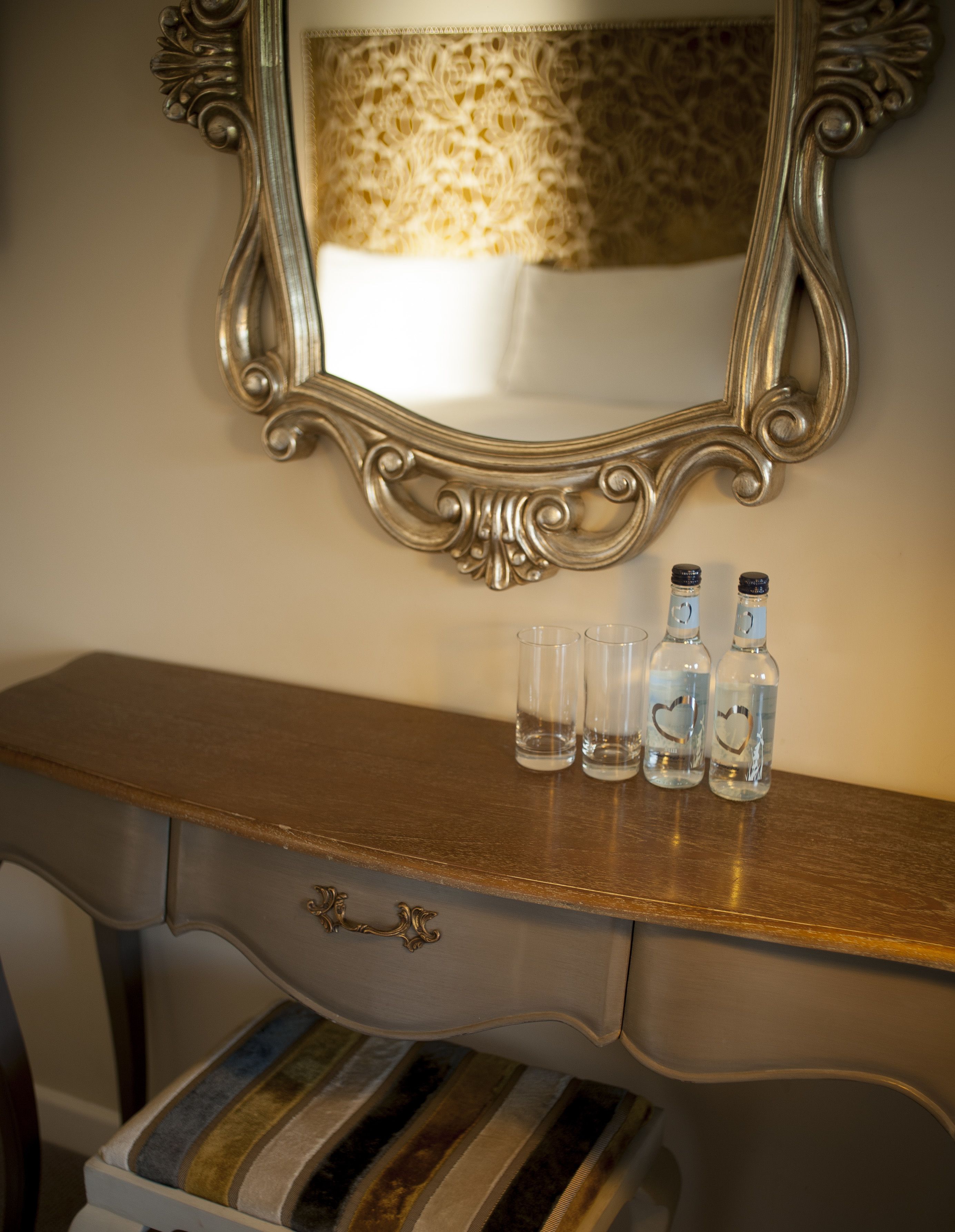 A vanity with a mirror, a wooden dresser, and three bottles on the dresser.
