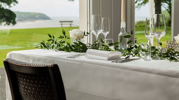 A dining table set with white tablecloth, glassware, and candles, overlooking a scenic view of a lake and greenery. - Mansion House Llansteffan (Carmarthen)