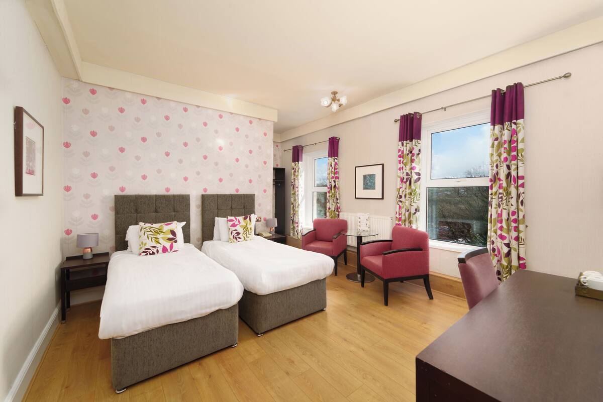 Habitación de hotel con dos camas, una mesita, una silla y vistas al exterior.