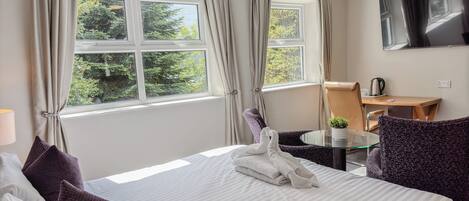 A hotel room with a bed, a small table with a kettle, a chair, and a TV on the wall.