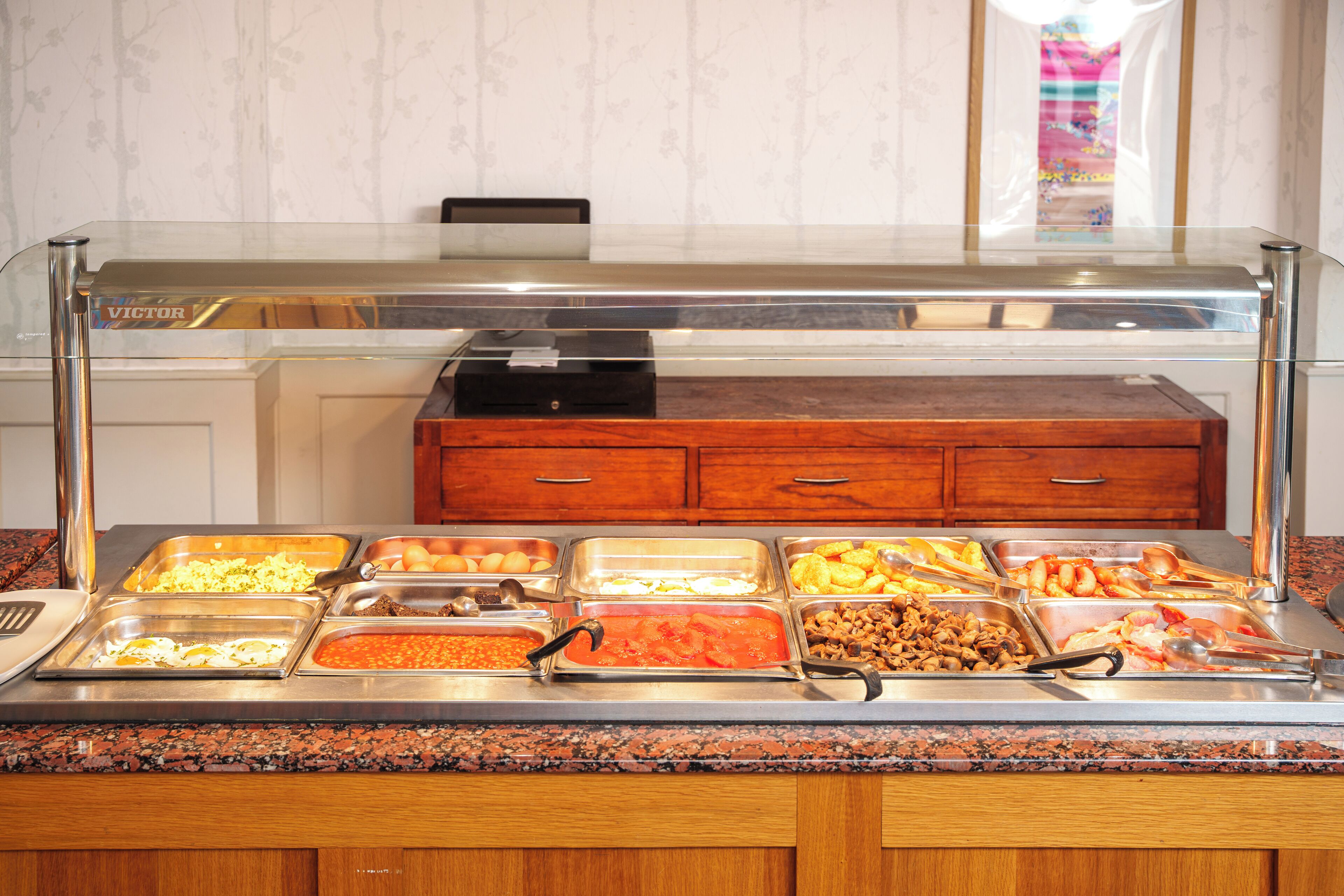 A buffet with various food items, a cash register, and wooden cabinets in the background.