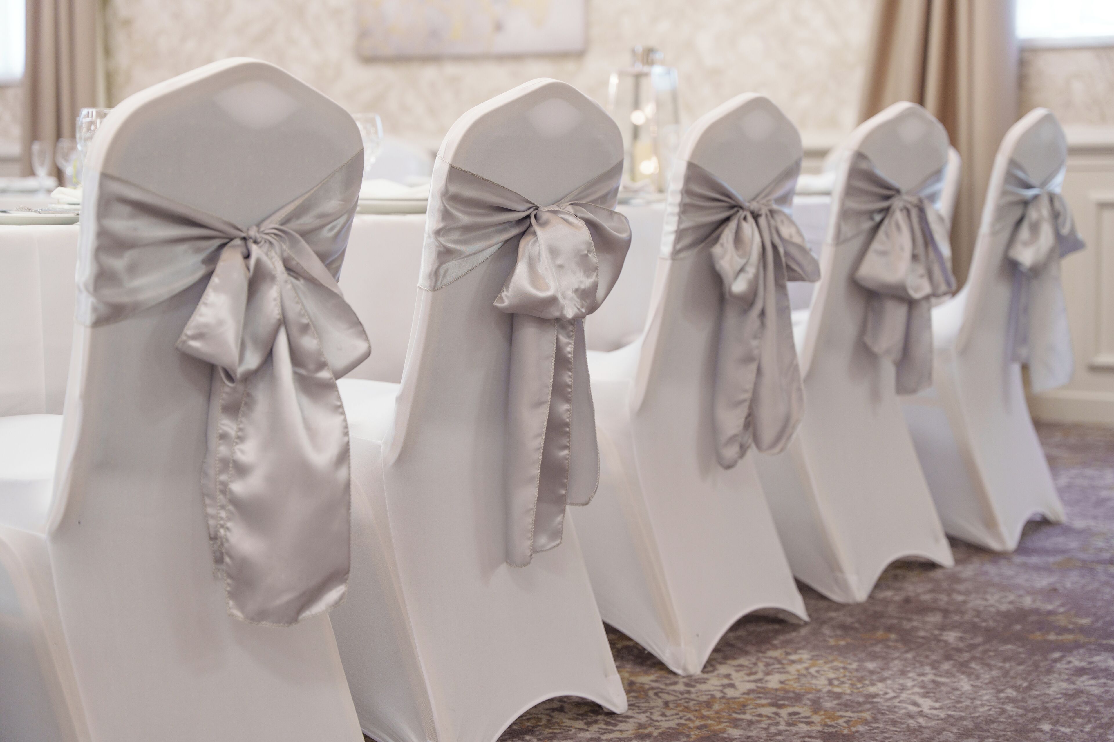 Rows of white chairs with grey satin bows, arranged in a banquet hall with a carpeted floor and a table set with glassware in the background.