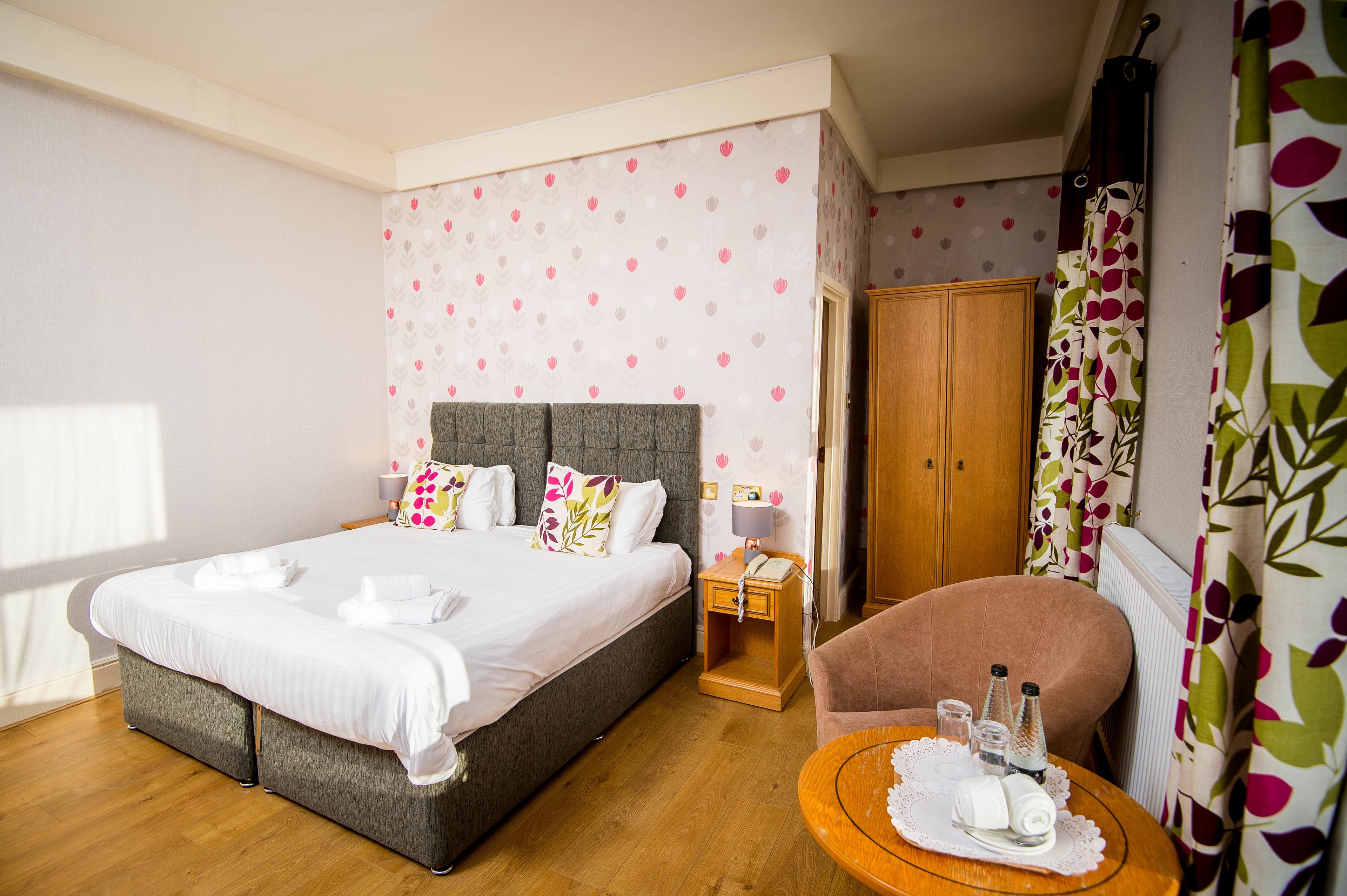 A hotel room with a large bed, a wooden wardrobe, a small table with toiletries, and a chair.