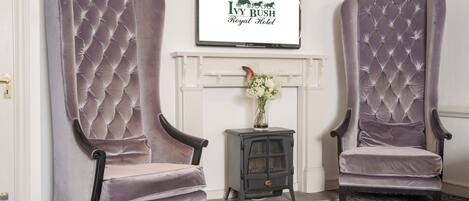 Two large, tufted armchairs in a room with a fireplace, a framed sign above, and a small table with a vase.