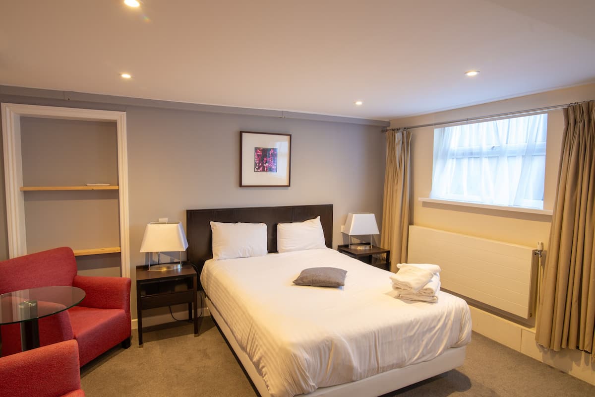 Una habitación de hotel con cama, un sillón rojo, una mesita de noche con lámpara y una ventana con cortinas.