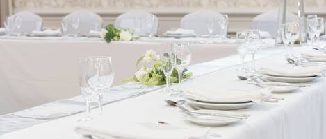 A long dining table set with white tablecloths, plates, glasses, and cutlery, with a floral centerpiece.