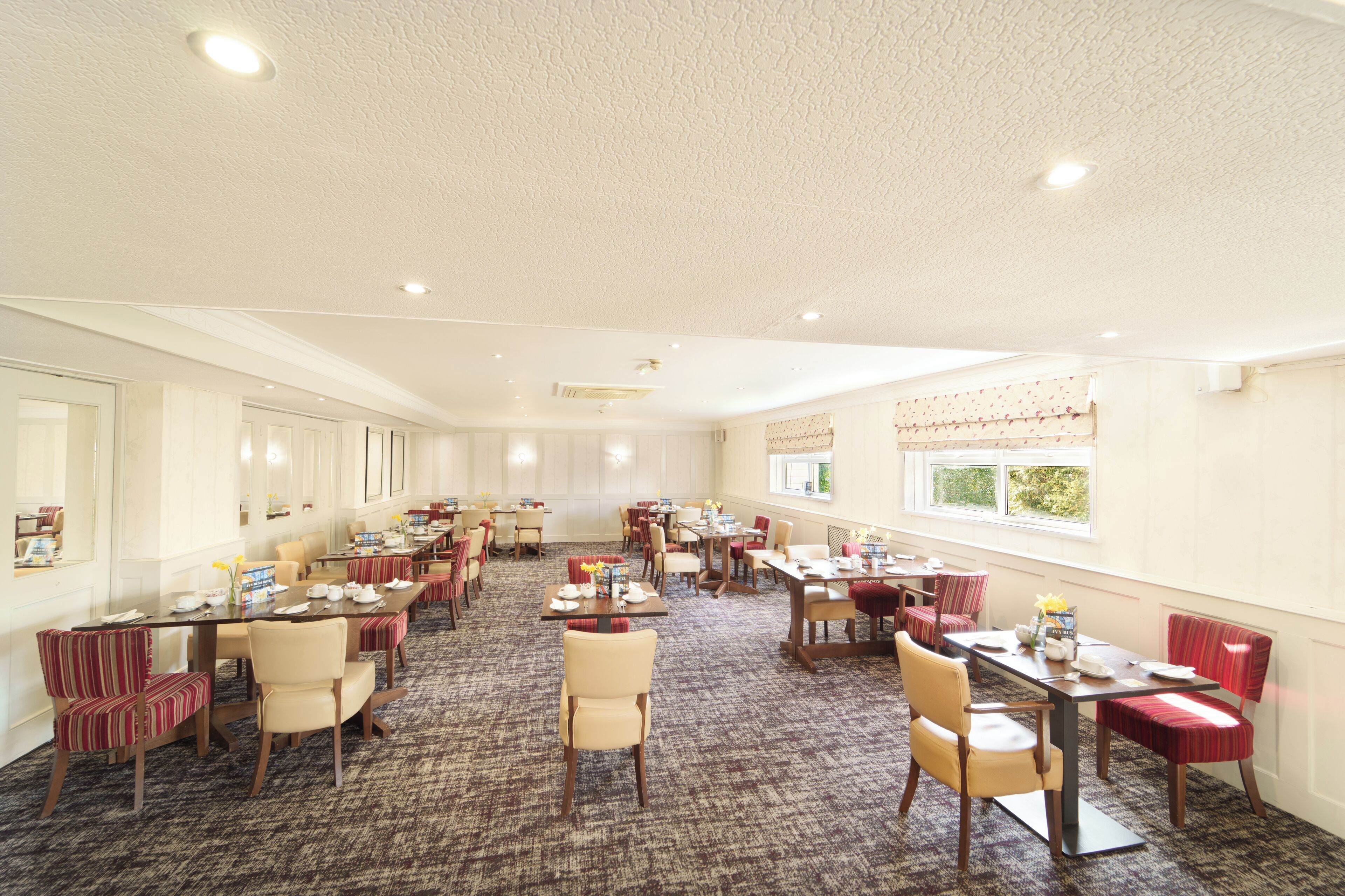 A spacious hotel dining area with multiple tables and chairs, large windows, and a carpeted floor.