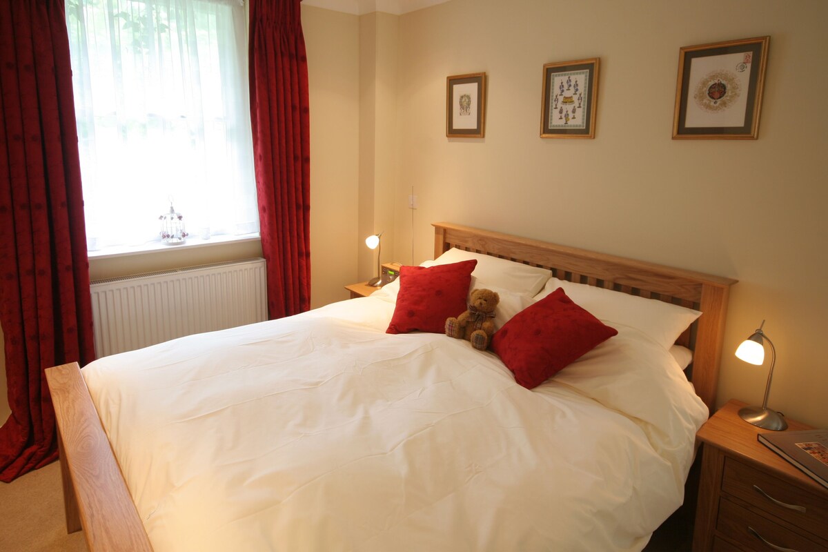 A neatly made bed with white linens and red pillows, a wooden headboard, and a nightstand with a lamp.