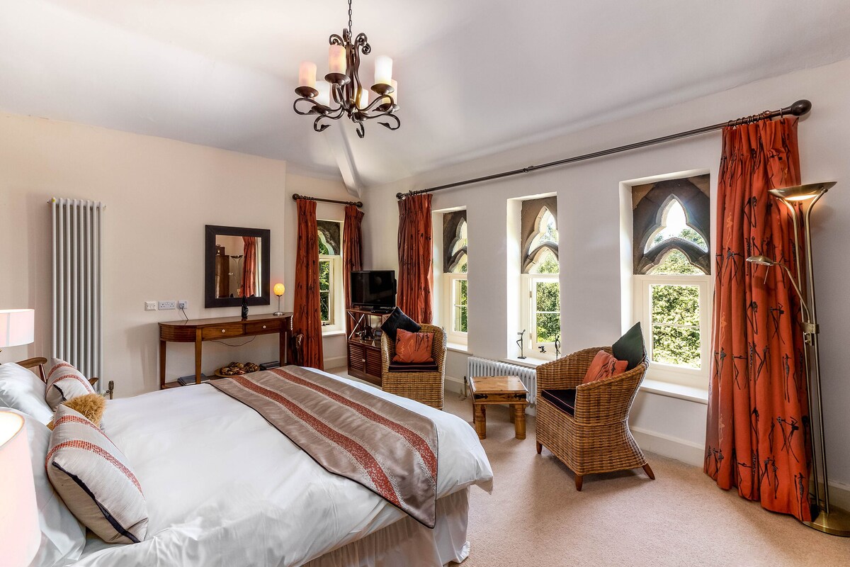 A bedroom with a large bed, a television, a desk, and a chair. There are windows with curtains, a chandelier, and a radiator.