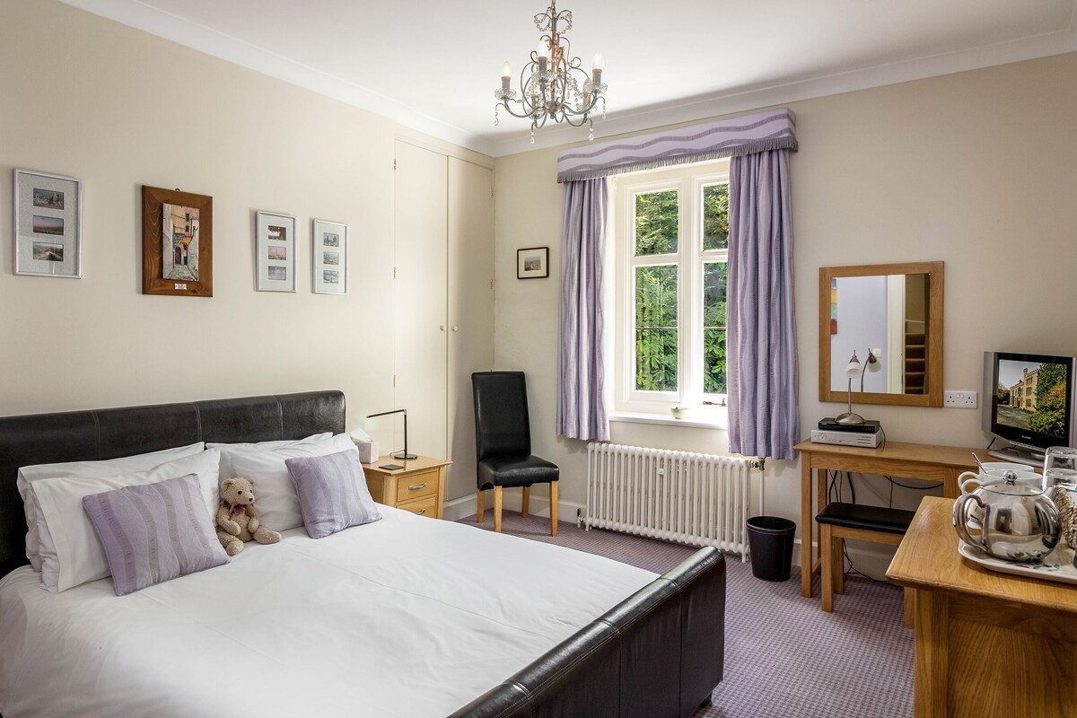 A bedroom with a large bed, a desk with a computer, a chair, and a window with curtains.