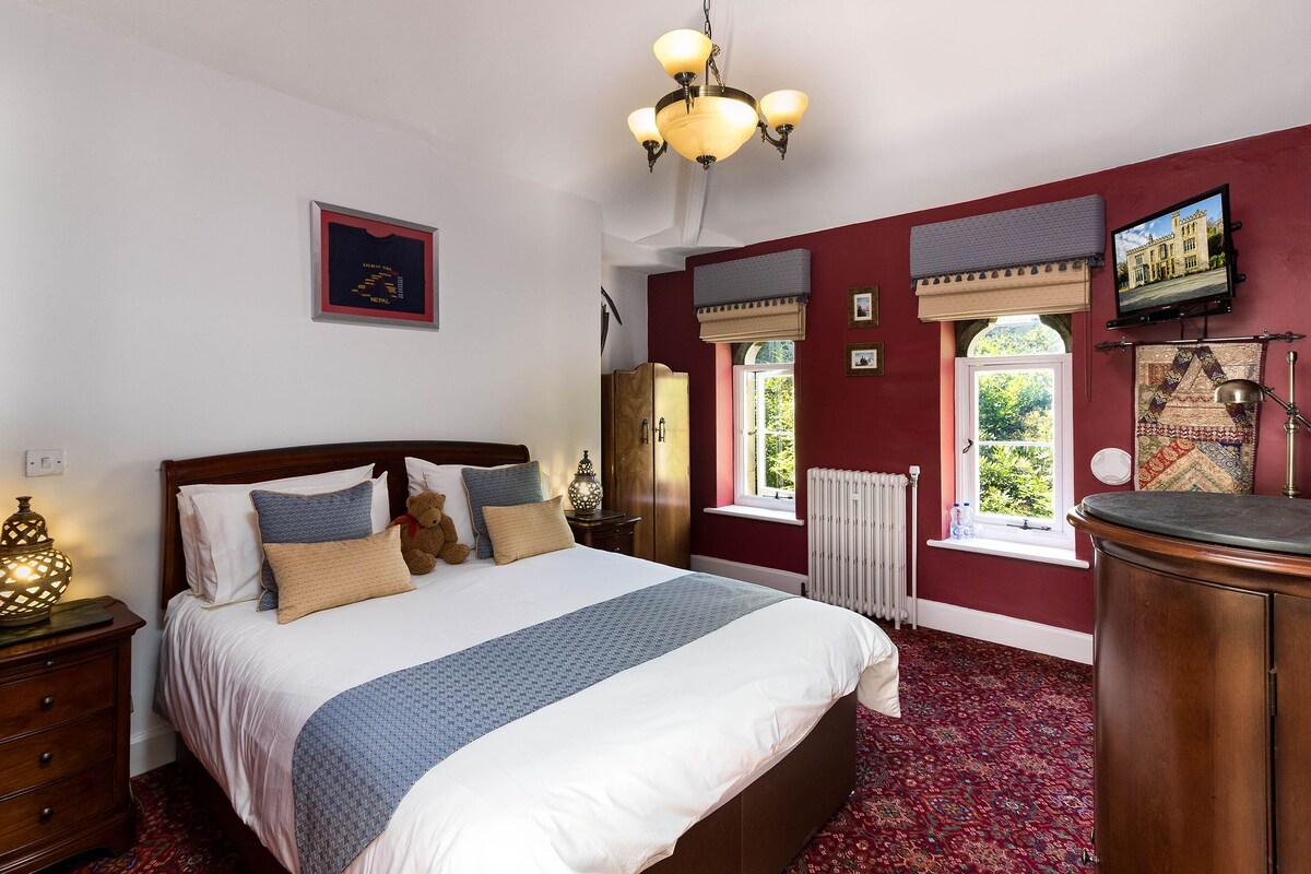 A bedroom with a large bed, a wooden nightstand, a dresser, a chandelier, and a window with a view of greenery.