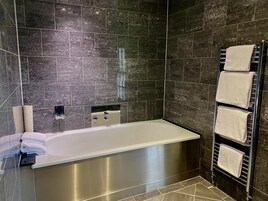 Un baño moderno con una bañera grande, paredes revestidas de baldosas oscuras y un perchero con toallas dobladas.