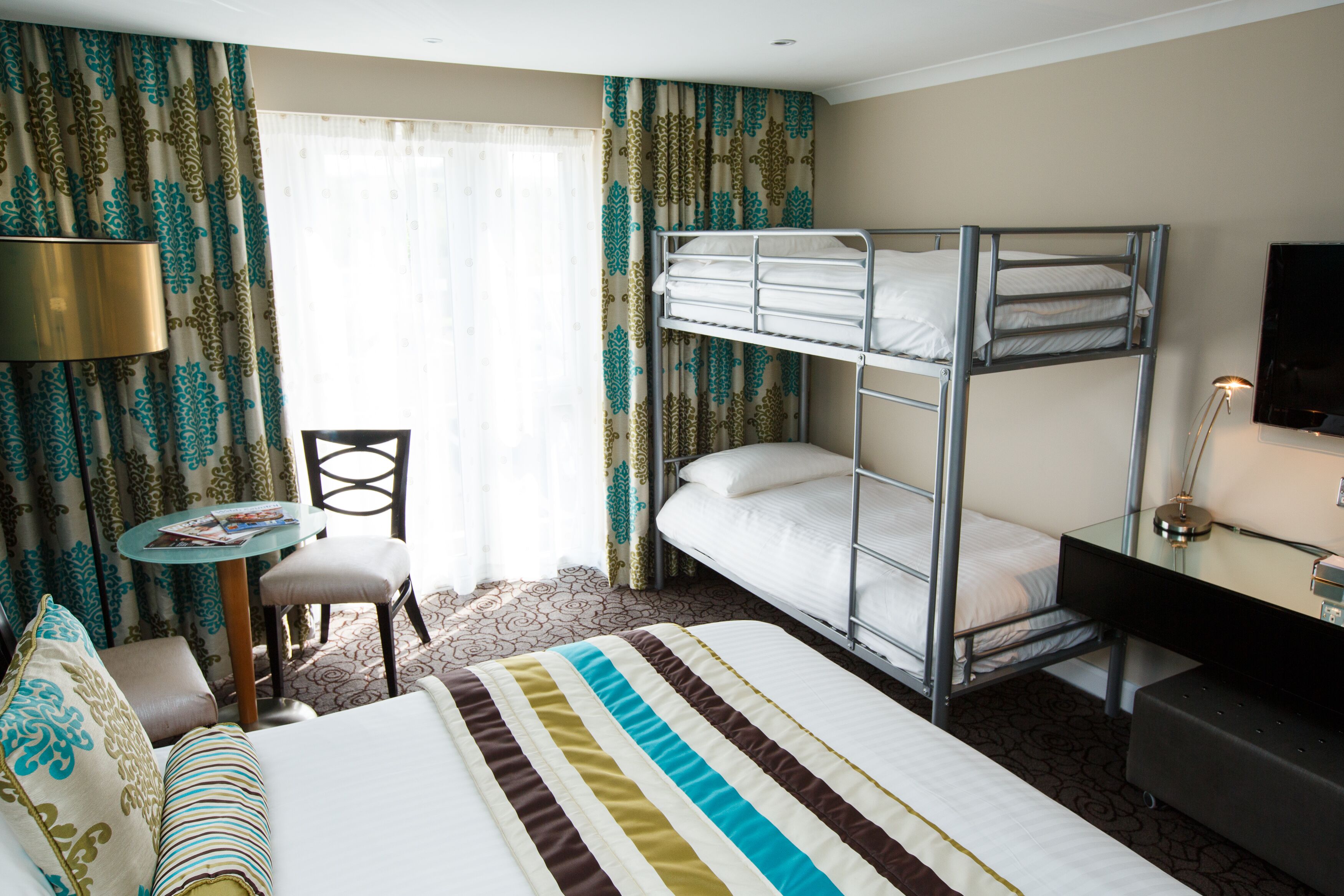 Una habitación de hotel con literas, un escritorio con lámpara, una silla y una pequeña mesa con revistas.