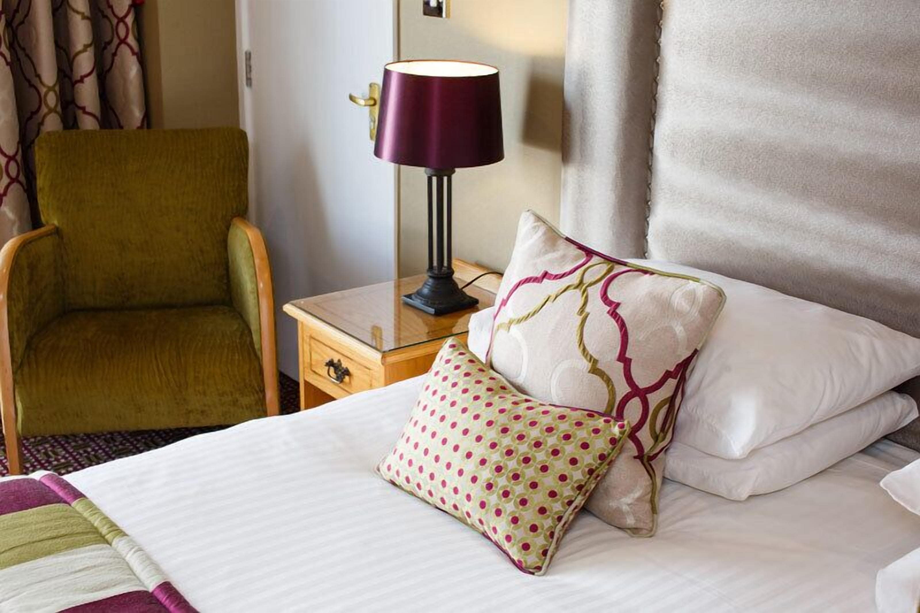 A hotel room with a bed, a green armchair, a nightstand with a lamp, and patterned pillows.