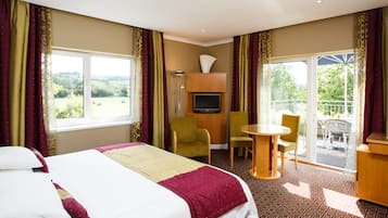 Habitación de hotel con cama, dos sillas, una mesita, televisión y balcón con vistas.