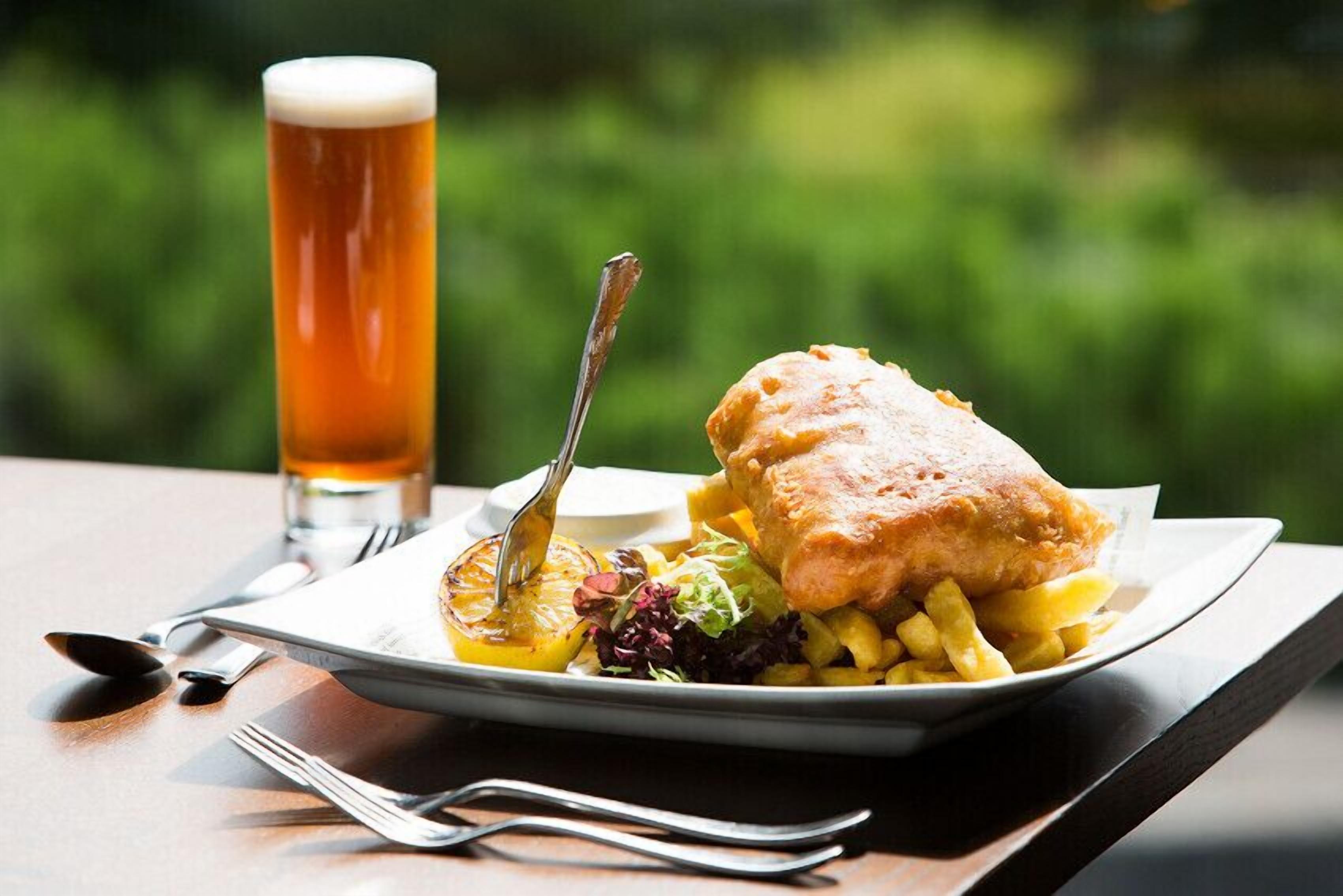 Un plato con pescado frito, papas fritas, una cerveza en un vaso y una ensalada como guarnición sobre una mesa con tenedor y cuchara.