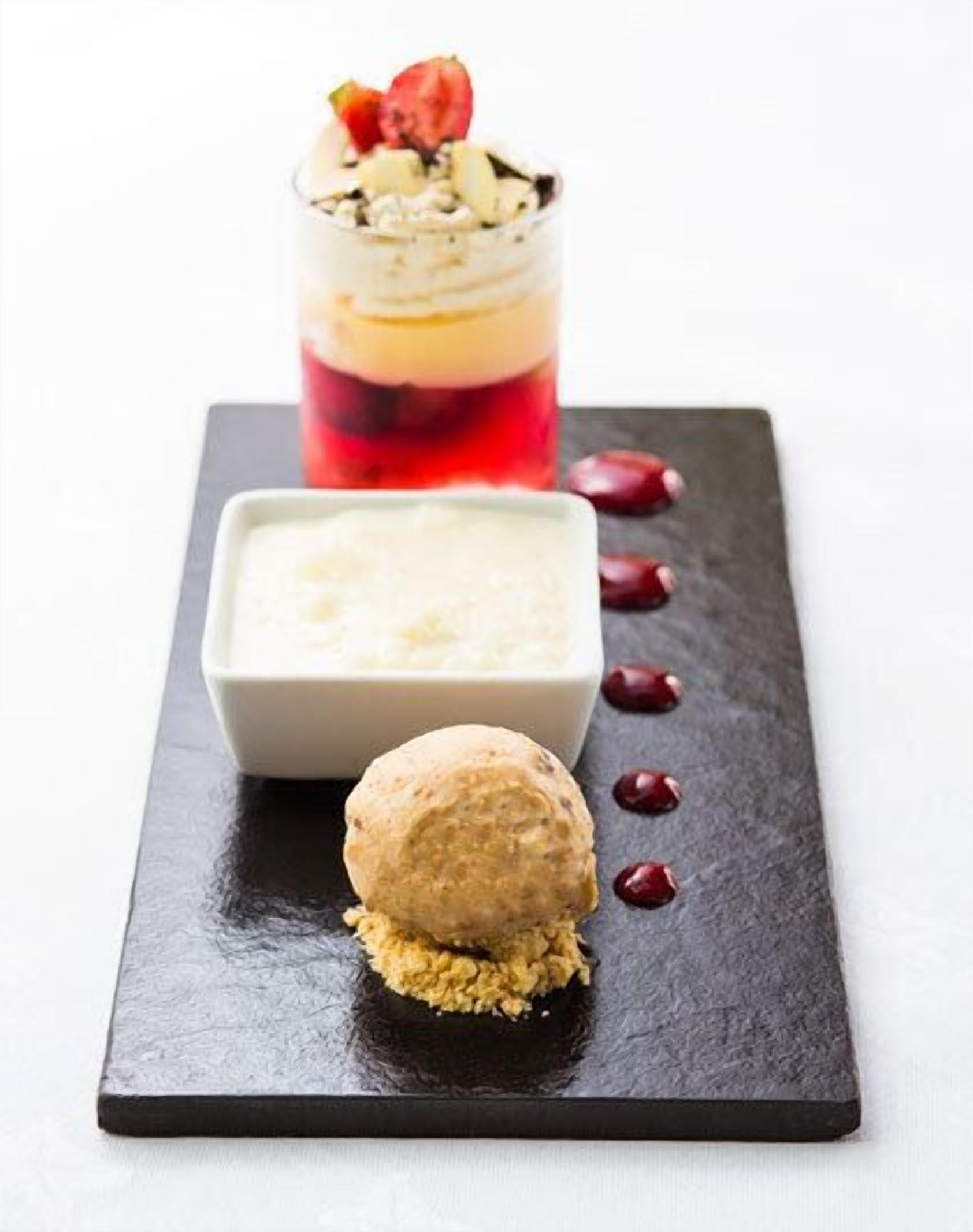 A dessert presentation on a slate board with a layered dessert in a glass, a square dish with a creamy dessert, and a round dessert with a biscuit base.