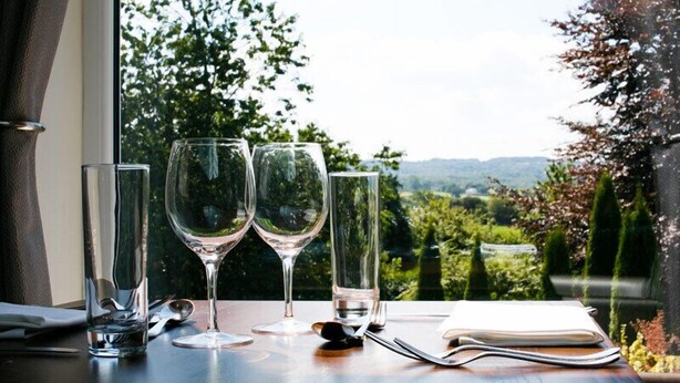 A table with three wine glasses and a water bottle, a notebook, and a pair of sunglasses, set against a backdrop of a lush green landscape visible through a window. - The Plough Rhosmaen (Llandeilo)