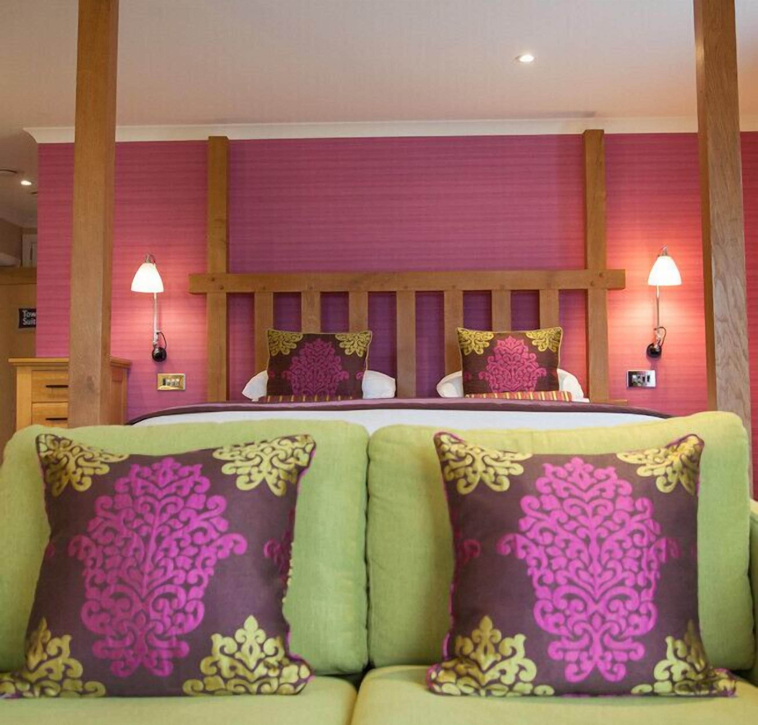 Habitación de hotel con una cama grande, dos lámparas de noche y sofás tapizados en verde con almohadones decorativos.