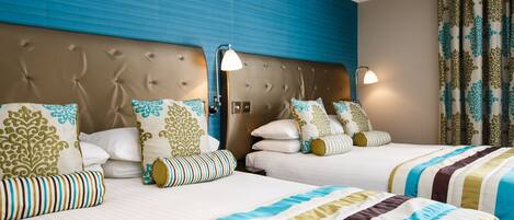 Habitación de hotel con dos camas, cada una con ropa de cama a rayas y almohadas decorativas. La habitación cuenta con un cabecero capitonado, una lámpara de pared y una cortina estampada.
