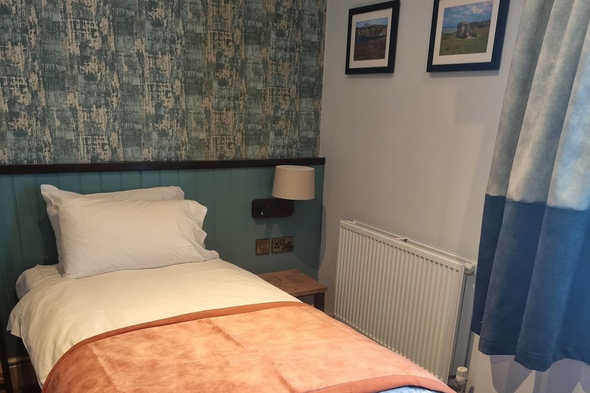 A neatly made bed with a grey mattress and an orange blanket, a white pillow, and a teal headboard. There is a small bedside table with a lamp, two framed pictures on the wall, and a window with a blue curtain.