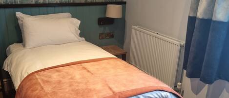 Una cama bien hecha con colchón gris y manta naranja, almohada blanca y cabecera color turquesa. Hay una mesita de noche con una lámpara, dos cuadros enmarcados en la pared y una ventana con cortina azul.