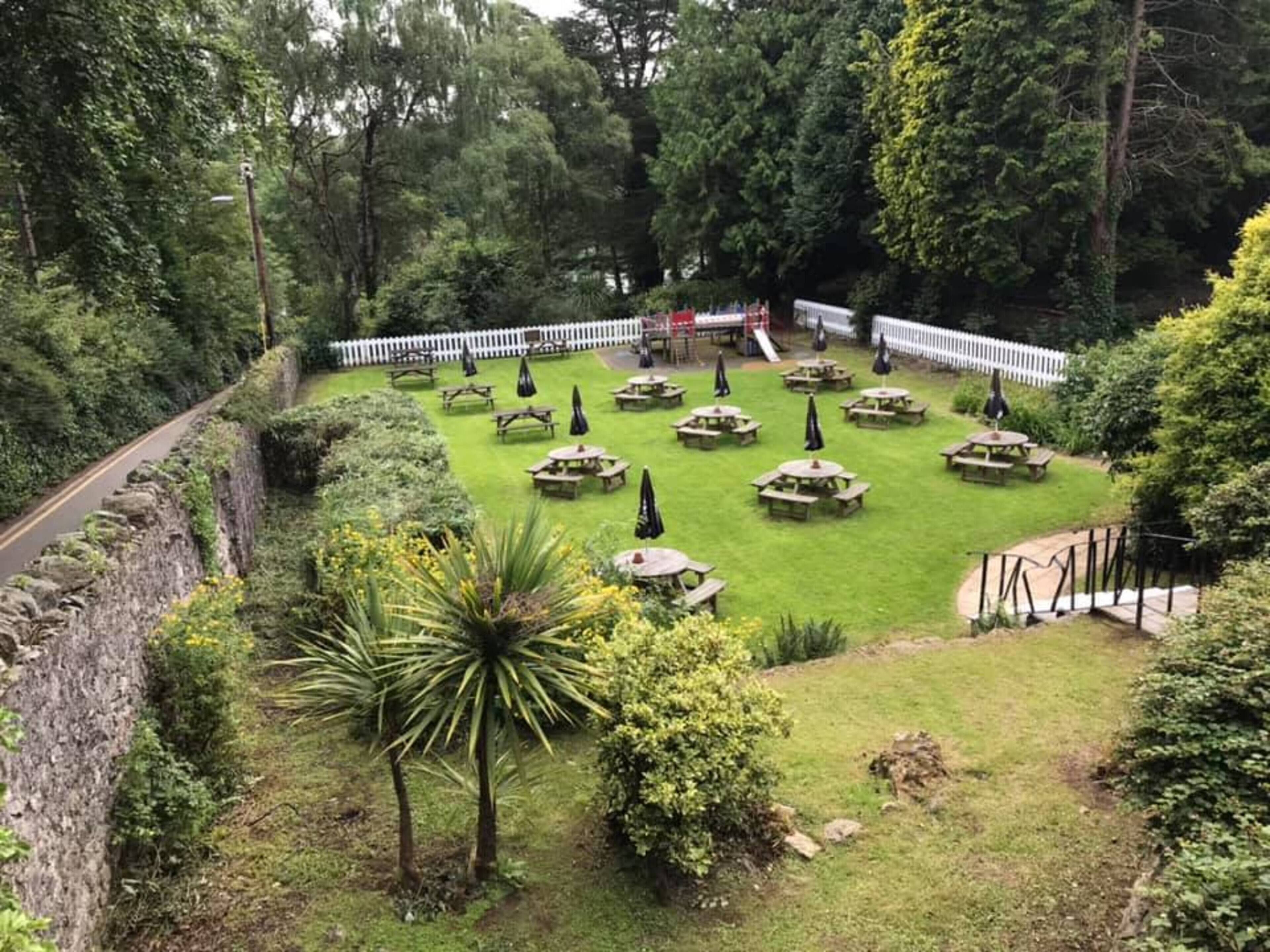 A well-maintained garden with picnic tables and umbrellas, surrounded by a white fence and dense greenery.
