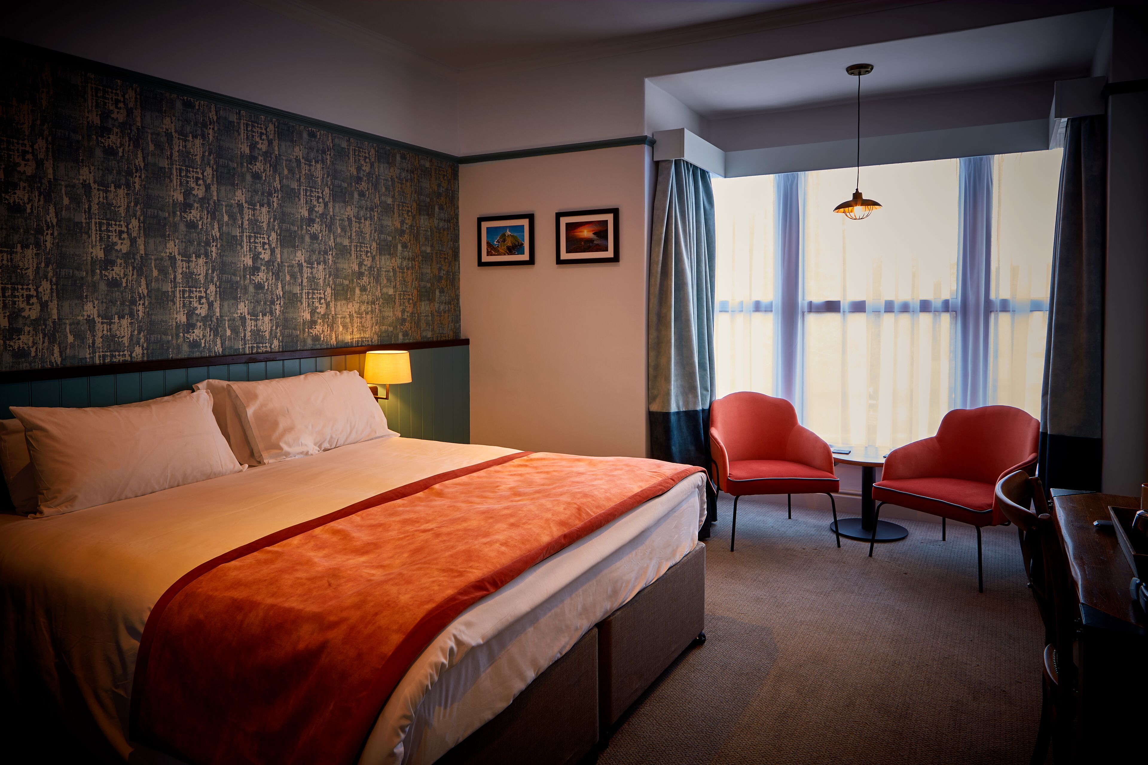 Habitación de hotel con una cama grande, dos sillas rojas, una mesita, una lámpara y vistas a la ciudad a través de amplios ventanales.