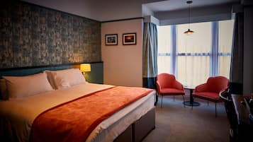 Ein Hotelzimmer mit einem großen Bett, zwei roten Sesseln, einem kleinen Tisch, einer Lampe und Blick auf die Stadt durch große Fenster.