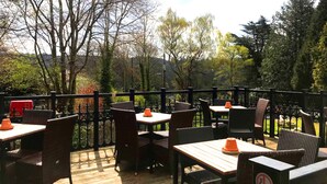 An outdoor patio with tables and chairs, surrounded by a wooden deck and metal railing, overlooking a garden with trees and a red vehicle in the background. - The Anglesey Arms (Menai Bridge)