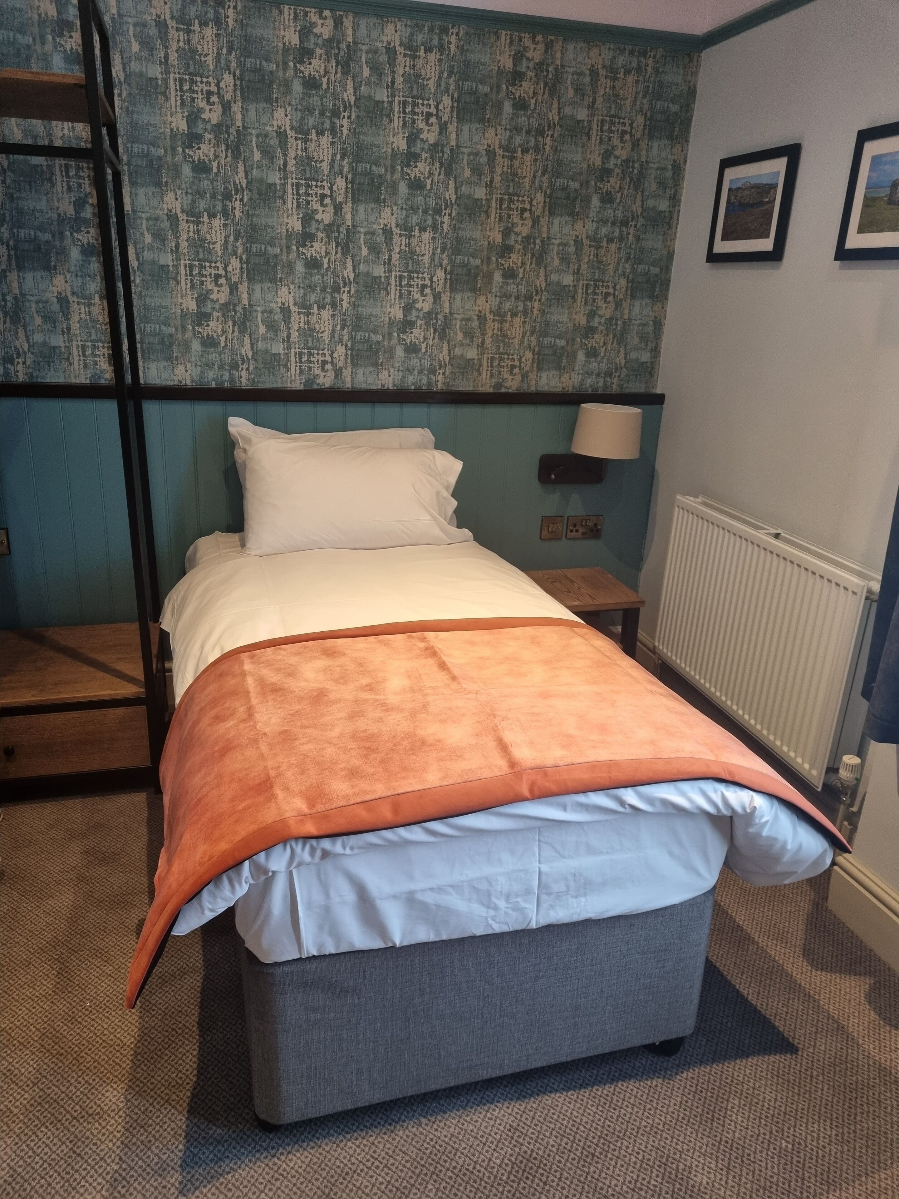 A neatly made bed with a grey mattress and an orange blanket, a wooden ladder, a small bedside table with a lamp, and a radiator.