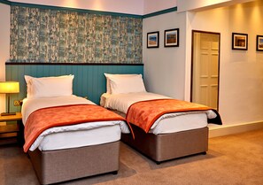 A hotel room with two beds, a nightstand, a lamp, and a door. - The Anglesey Arms (Menai Bridge)