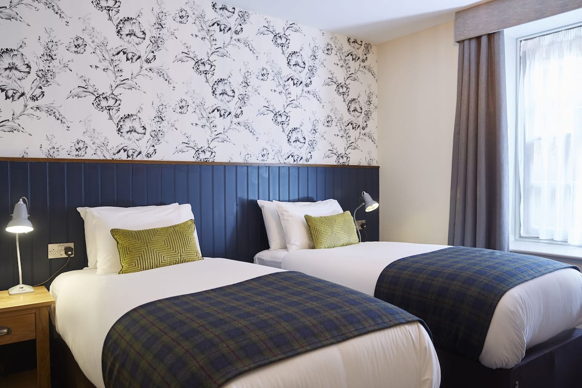A hotel room with two beds, a floral wallpaper, a wooden headboard, and a window with curtains.