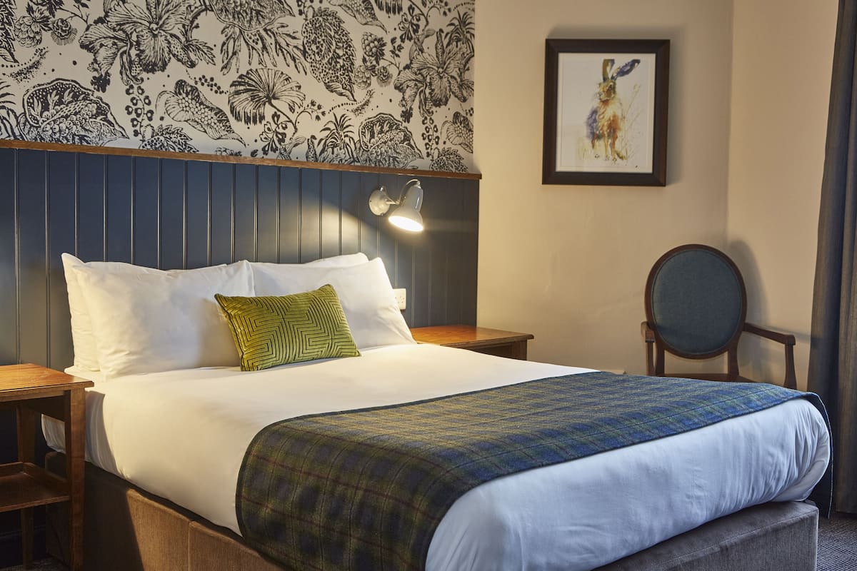 A neatly made bed with white linens and a plaid blanket, a wooden nightstand, a green pillow, a wall-mounted lamp, and a framed picture on the wall.
