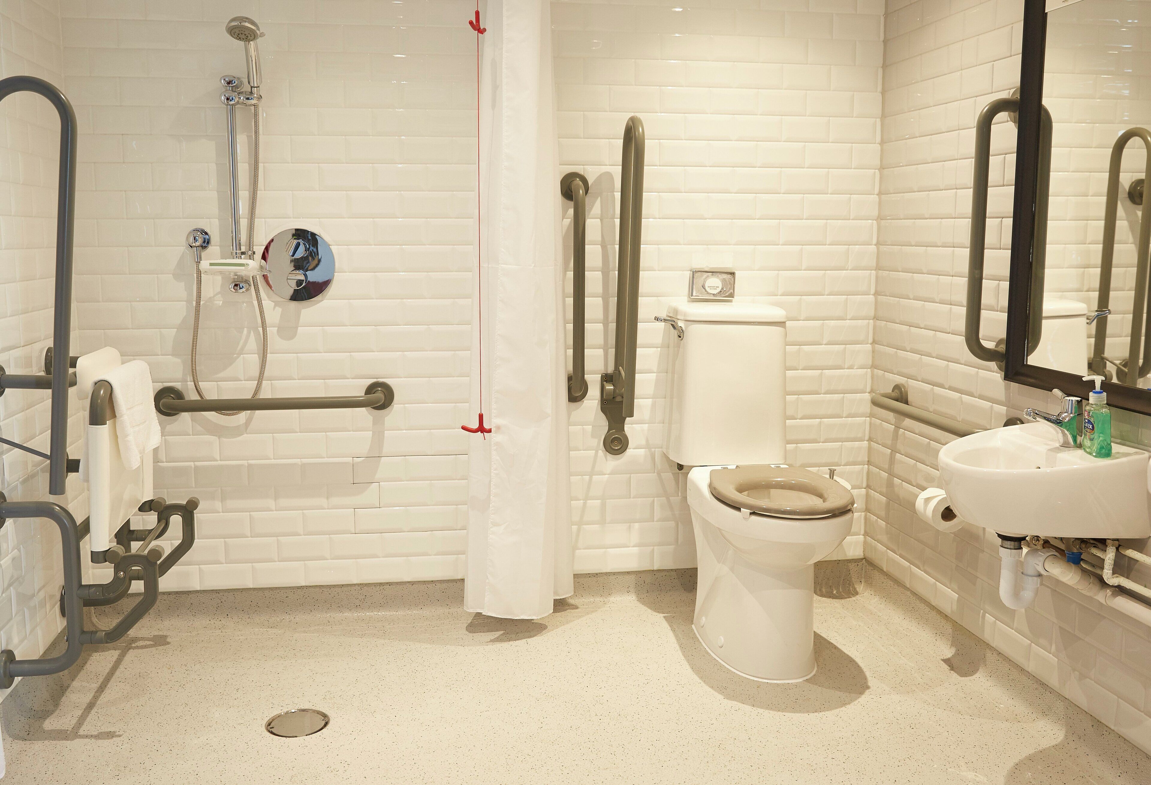 Un baño con paredes de azulejos blancos, inodoro blanco, lavamanos y zona de ducha con barras de apoyo.