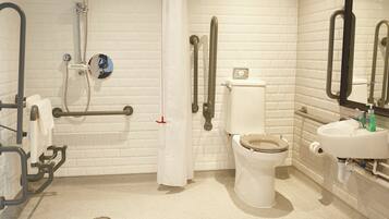 Un baño con paredes de azulejos blancos, inodoro blanco, lavamanos y zona de ducha con barras de apoyo.