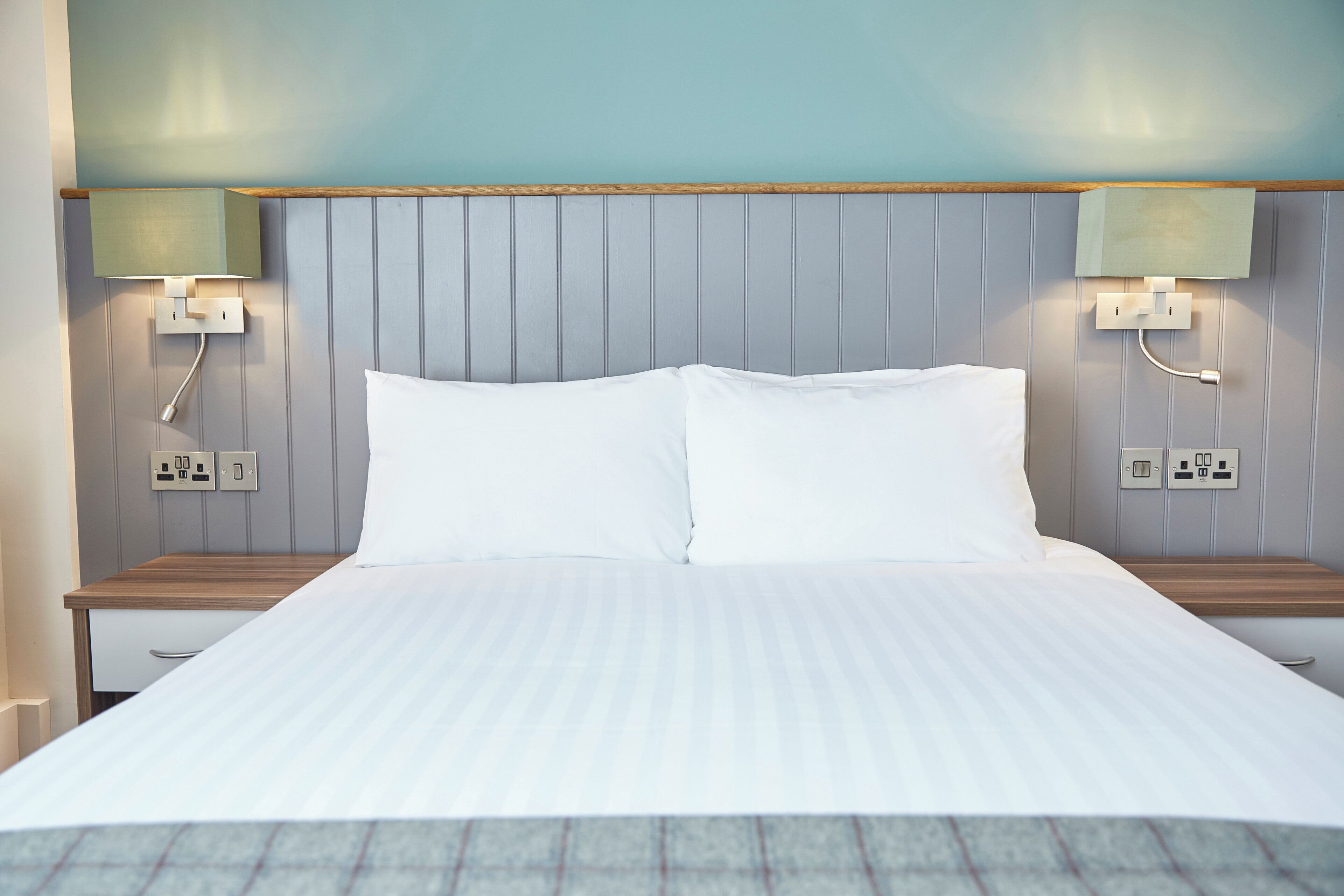 A neatly made bed with white linens, a wooden bedside table, and a wall-mounted nightstand with a lamp.