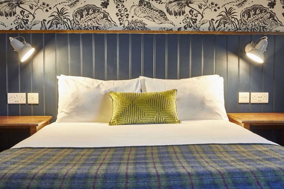 Een bed met witte lakens, een kussen met een groen patroon en een geruit plaid. Twee bedlampen en een muur met verticale houten panelen en bloemenbehang.