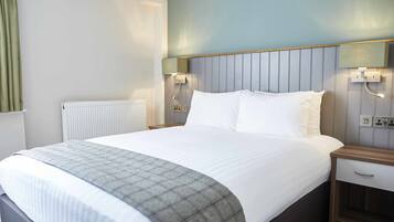 A hotel room with a large bed, a bedside table, a nightstand, a wall-mounted lamp, and a radiator.