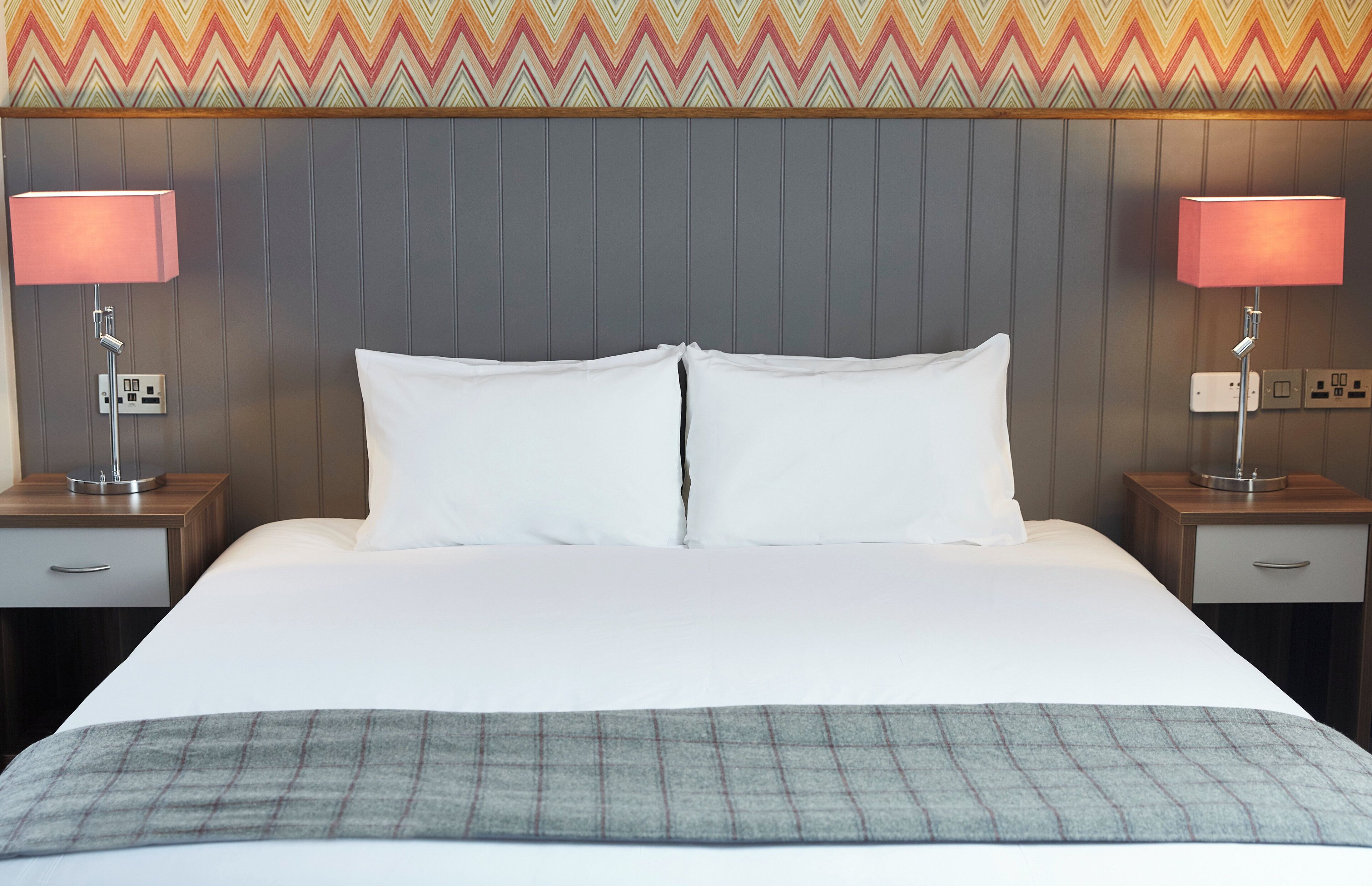A hotel room with a large bed, two bedside tables with lamps, a patterned wall, and a grey blanket.
