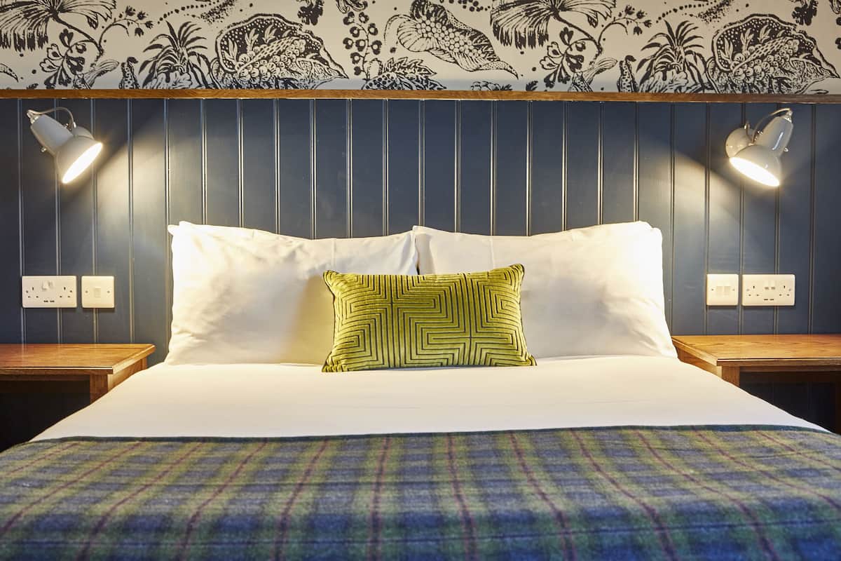 A neatly made bed with white and green pillows, a plaid blanket, and a wall with vertical paneling and floral wallpaper.