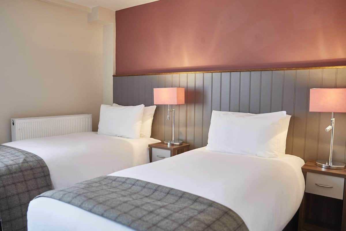 A hotel room with two beds, a radiator, a wooden headboard, and two bedside lamps.