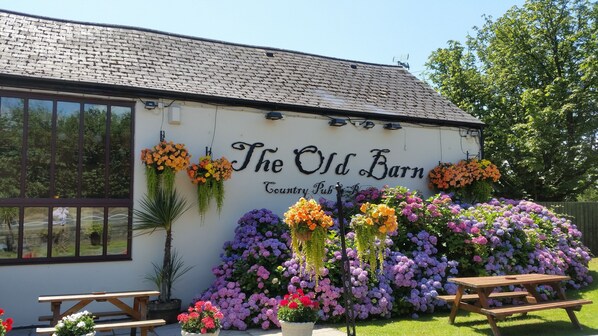 A white building with a sign reading "The Old Barn Country Pub." There are hanging flower baskets and a wooden bench outside.