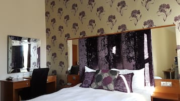 Habitación de hotel con cama, escritorio, silla, espejo y una pared con estampado floral.