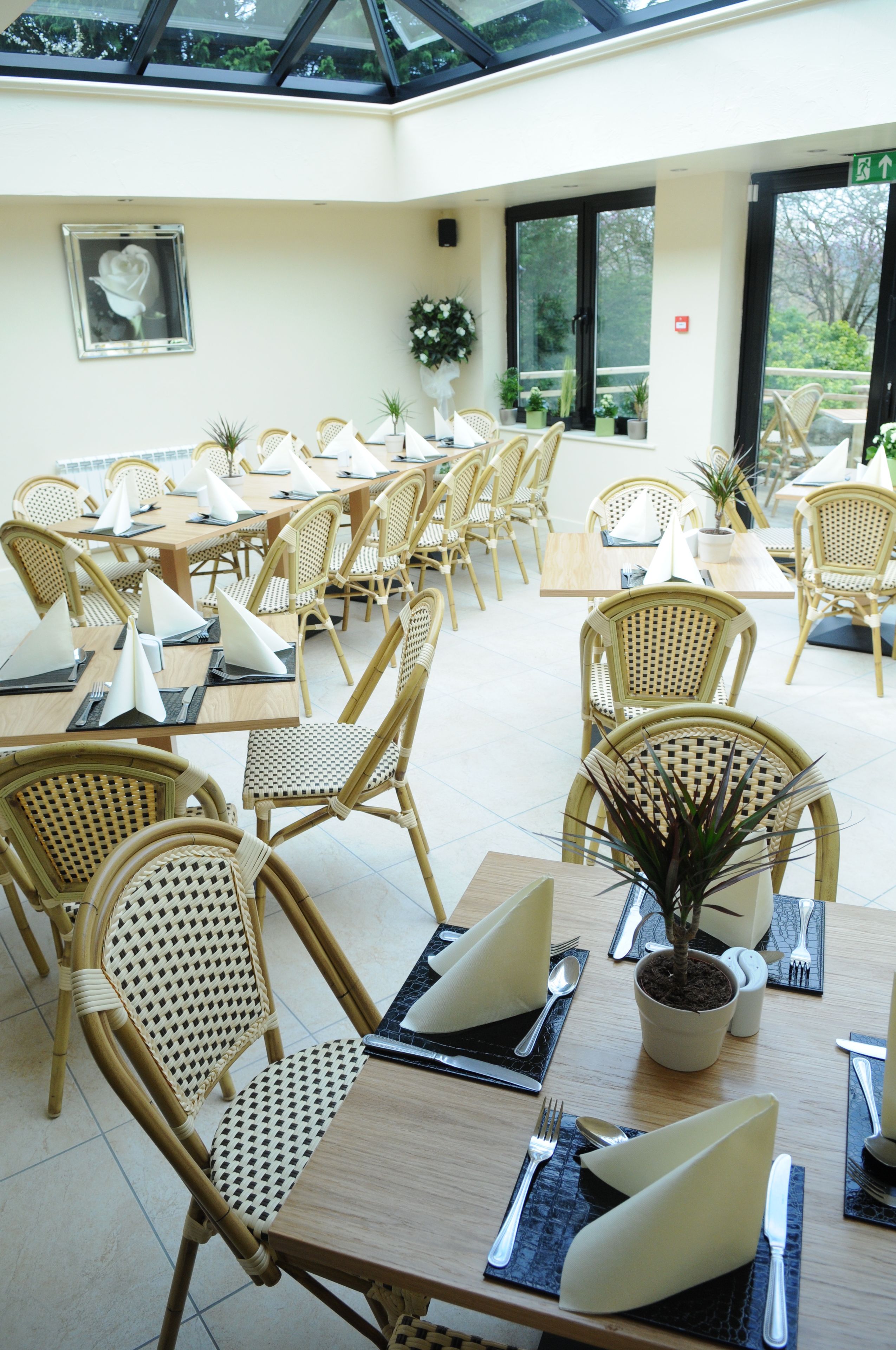 A spacious indoor dining area with multiple tables and wicker chairs, featuring a potted plant and a framed picture on the wall.