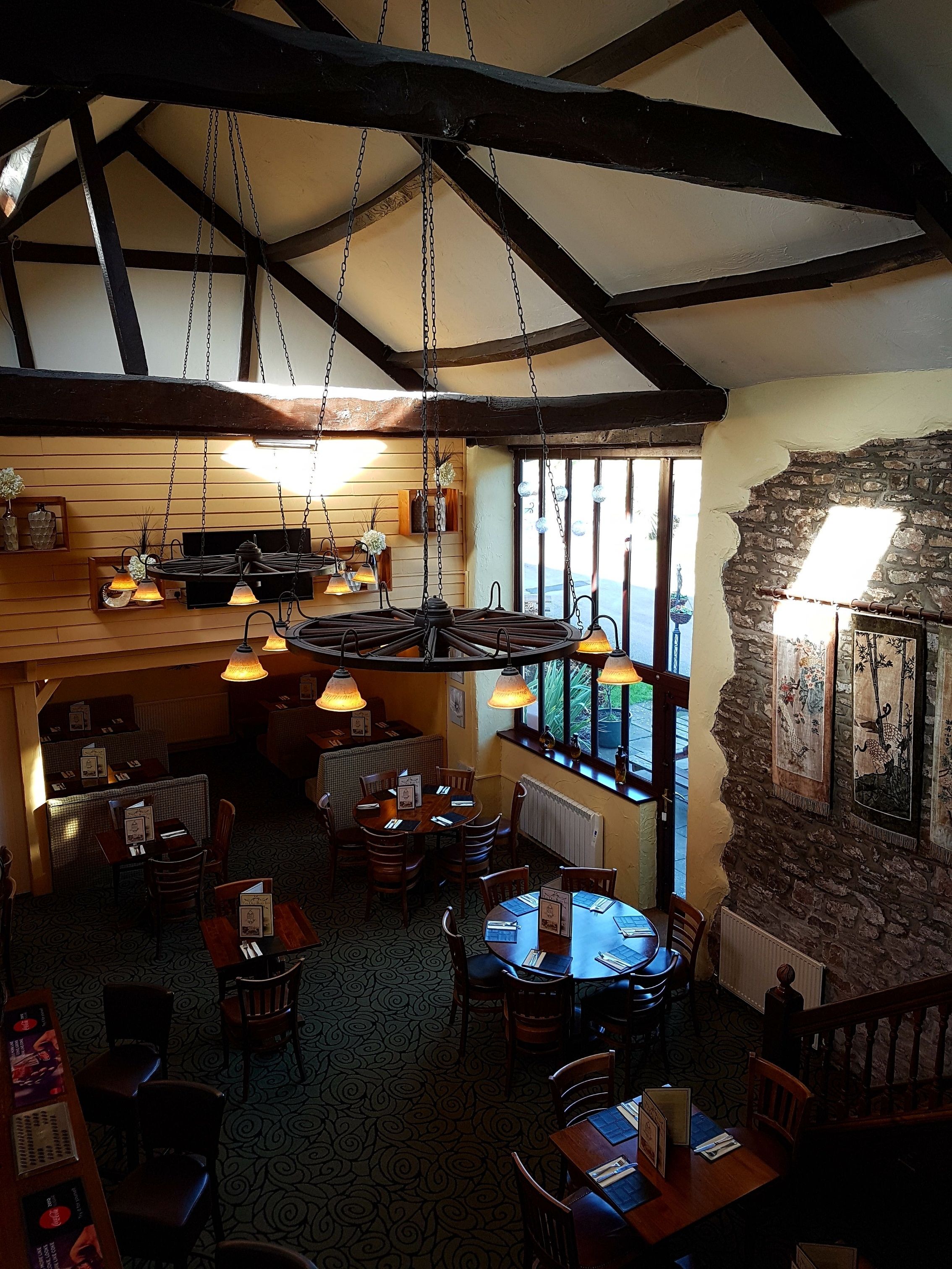 A restaurant with exposed wooden beams, hanging pendant lights, and a stone wall. Tables and chairs are arranged for dining.