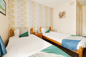 A double room with two beds, a wooden headboard, a nightstand with a lamp, and a wall with a leaf-patterned wallpaper.
