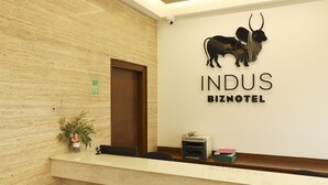 A reception desk with a logo for 'INDUS BIZNOTEL' and a bull sculpture. There is a door, a plant, and a printer on the desk. - Indus Biznotel (New Delhi)